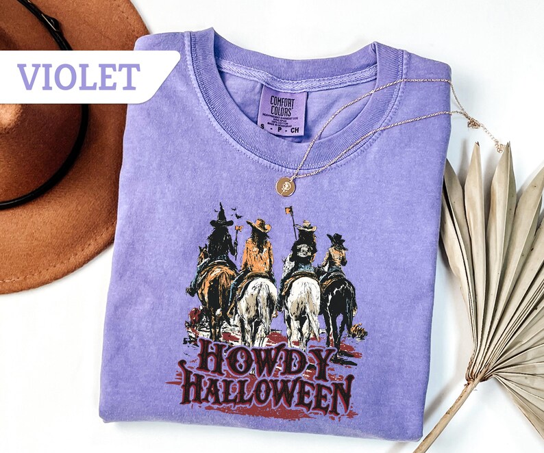 May include: Violet Comfort Colors t-shirt with a graphic of four cowgirls on horseback, and the text "HOWDY HALLOWEEN". A gold necklace and a brown hat are also visible. The t-shirt is a relaxed fit.