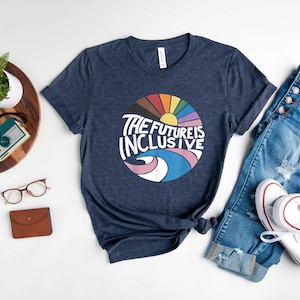 May include: A navy blue t-shirt with a graphic of a rainbow sun and the text "The Future is Inclusive". The shirt is laid out on a white surface with a pair of jeans, a watch, a camera, glasses, and a wallet.