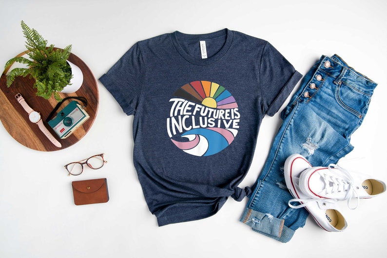 May include: A navy blue t-shirt with a graphic of a rainbow sun and the text "The Future is Inclusive". The shirt is laid out on a white surface with a pair of jeans, a watch, a camera, glasses, and a wallet.