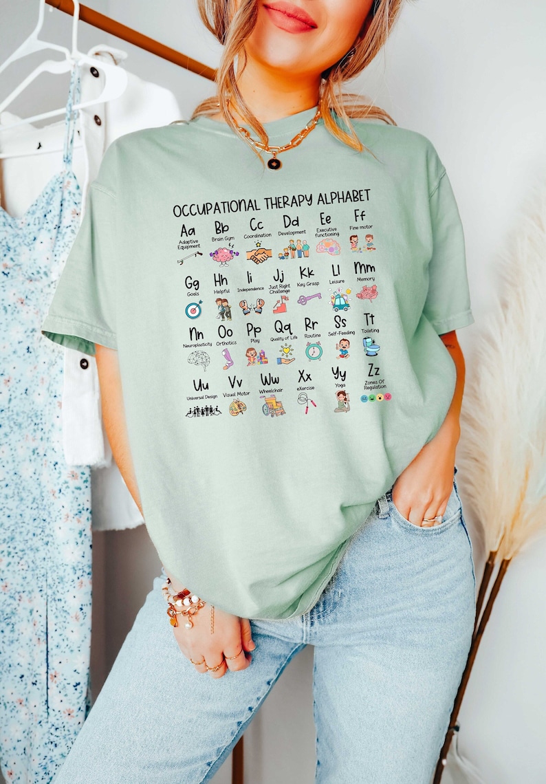May include: A light green t-shirt with the text "Occupational Therapy Alphabet" and an alphabet chart with icons representing different occupational therapy concepts. The icons include a brain, a hand shake, a key, a target, a wheelchair, and a sun.