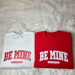 May include: Two folded sweatshirts, one white and one red. Each features the text "BE MINE" in a bold, collegiate-style font, with "VALENTINE" underneath. The white sweatshirt has red lettering, while the red sweatshirt has white lettering.