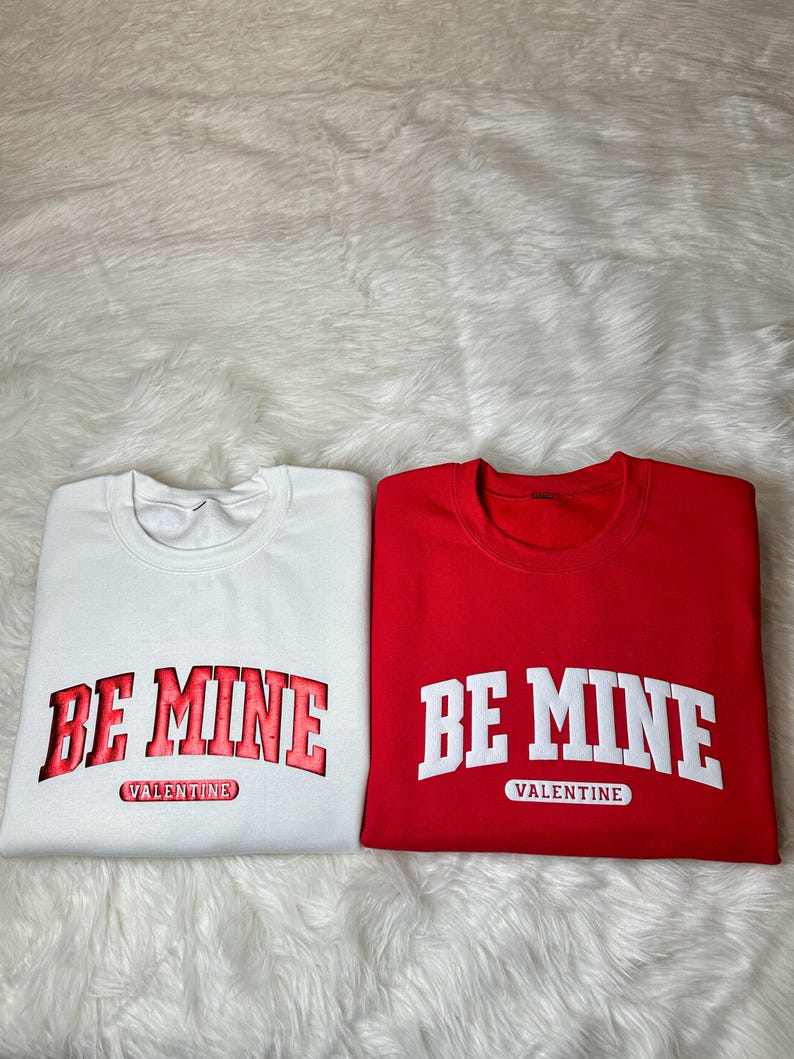 May include: Two folded sweatshirts, one white and one red. Each features the text "BE MINE" in a bold, collegiate-style font, with "VALENTINE" underneath. The white sweatshirt has red lettering, while the red sweatshirt has white lettering.