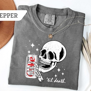 May include: Dark gray t-shirt featuring a white skull holding a Diet Coke can. The design includes the phrase "'til death." The shirt is adorned with a delicate necklace. The shirt is a Comfort Colors brand.