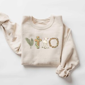 May include: A beige sweatshirt with a graphic design of a cross, a lamb, and a crown of thorns. The design is in shades of brown, green, and white.