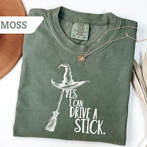 May include: Moss green t-shirt with a witch hat and broom graphic. The text reads "Yes I Can Drive A Stick." A gold necklace with a pendant is on the shirt. A brown hat and dried palm leaves are in the background.