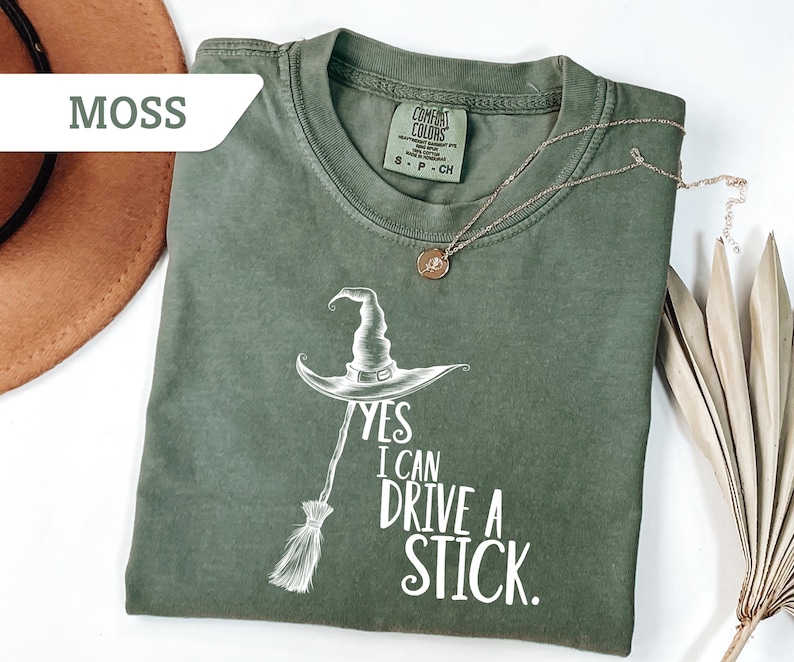 May include: Moss green t-shirt with a witch hat and broom graphic. The text reads "Yes I Can Drive A Stick." A gold necklace with a pendant is on the shirt. A brown hat and dried palm leaves are in the background.