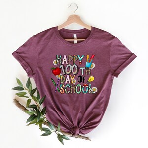 Puede incluir: Una camiseta color granate con el texto "Happy 100th Day of School" en letras de colores decoradas con &uacute;tiles escolares y flores.