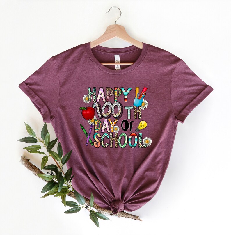 Puede incluir: Una camiseta color granate con el texto "Happy 100th Day of School" en letras de colores decoradas con &uacute;tiles escolares y flores.
