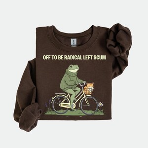 May include: A dark brown sweatshirt with the text "OFF TO BE RADICAL LEFT SCUM". The graphic shows a frog riding a bicycle with a cat in the basket. The frog wears a green sweater.