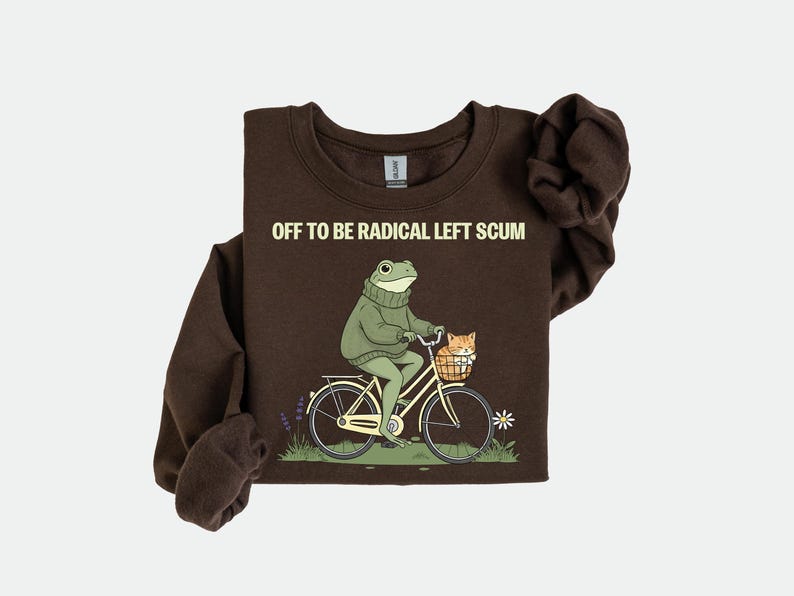 May include: A dark brown sweatshirt with the text "OFF TO BE RADICAL LEFT SCUM". The graphic shows a frog riding a bicycle with a cat in the basket. The frog wears a green sweater.