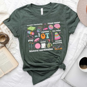 May include: A dark green t-shirt with a colorful graphic design featuring the word "OT" surrounded by illustrations of various activities and skills related to occupational therapy, including play, social interaction, sensory processing, fine motor, visual motor, cognition, and bilateral coordination.