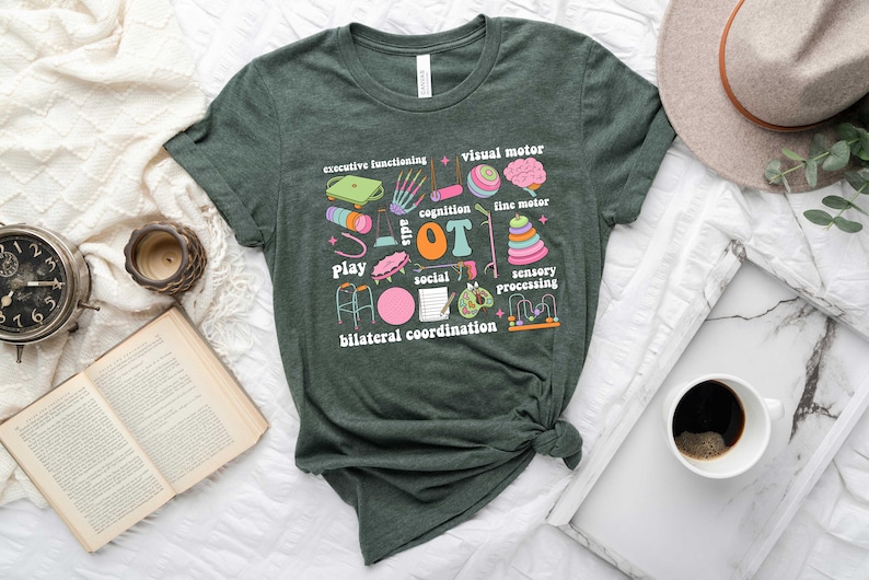 May include: A dark green t-shirt with a colorful graphic design featuring the word "OT" surrounded by illustrations of various activities and skills related to occupational therapy, including play, social interaction, sensory processing, fine motor, visual motor, cognition, and bilateral coordination.