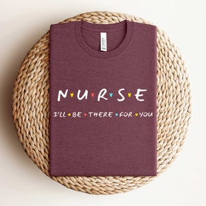 Puede incluir: Camiseta color burdeos con el texto "NURSE" en letras de colores con corazones entre cada letra. El texto "I'LL BE THERE FOR YOU" est&aacute; debajo de la palabra "NURSE" en letras negras con puntos de colores entre cada palabra.