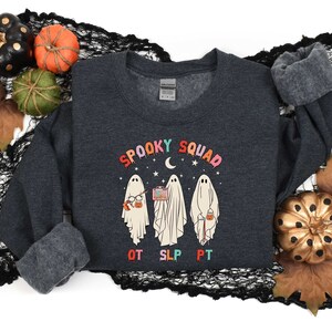 May include: A dark gray sweatshirt with a graphic of three ghosts wearing glasses, holding a book, a microphone, and a bucket of candy. The text "Spooky Squad" is above the ghosts, and "OT SLP PT" is below. The sweatshirt is on a black and white background with fall leaves and pumpkins.
