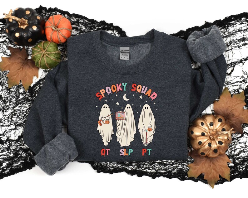 May include: A dark gray sweatshirt with a graphic of three ghosts wearing glasses, holding a book, a microphone, and a bucket of candy. The text "Spooky Squad" is above the ghosts, and "OT SLP PT" is below. The sweatshirt is on a black and white background with fall leaves and pumpkins.