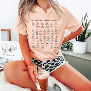 May include: A pink t-shirt with the text "Occupational Therapy Alphabet" and a grid of letters with corresponding icons and descriptions. The letters are A through Z, and the icons represent different aspects of occupational therapy.