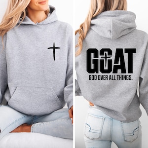 Pode incluir: Um moletom cinza claro com um bolso frontal e capuz com cord&atilde;o. A frente apresenta uma cruz preta, e as costas exibem a palavra "GOAT" com uma cruz no "O", e o texto "GOD OVER ALL THINGS."