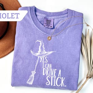 May include: A violet-colored t-shirt with the text "YES I CAN DRIVE A STICK" and a witch hat and broom graphic. The shirt has a crew neck and short sleeves. A gold necklace with a pendant is draped across the shirt.