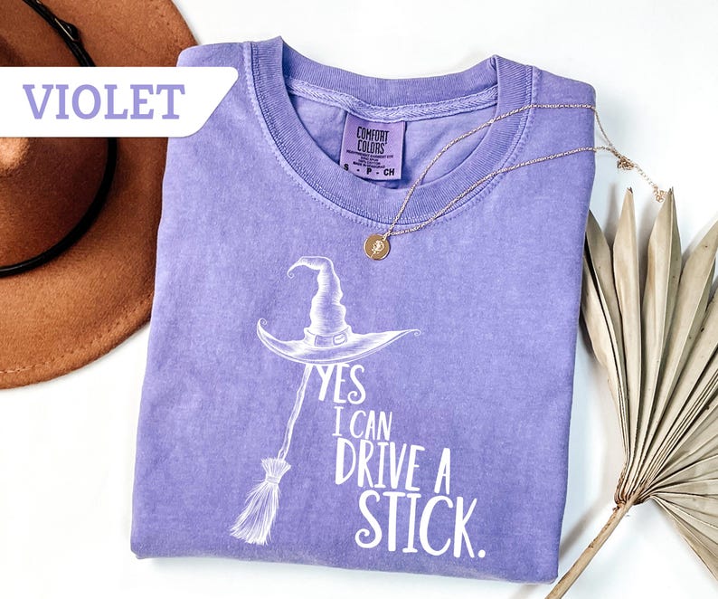 May include: A violet-colored t-shirt with the text "YES I CAN DRIVE A STICK" and a witch hat and broom graphic. The shirt has a crew neck and short sleeves. A gold necklace with a pendant is draped across the shirt.