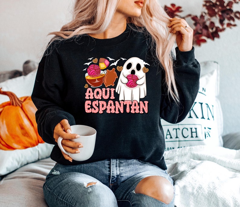 May include: A black sweatshirt featuring a ghost holding a pink pastry and the text "Aqui Espantan" in a retro font. The ghost is surrounded by bats and other Halloween-themed imagery.