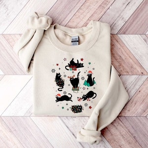 May include: A cream-coloured sweatshirt with a festive design featuring black cats wearing Santa hats, reindeer antlers, and Christmas lights. The cats are surrounded by snowflakes, stars, and Christmas trees.