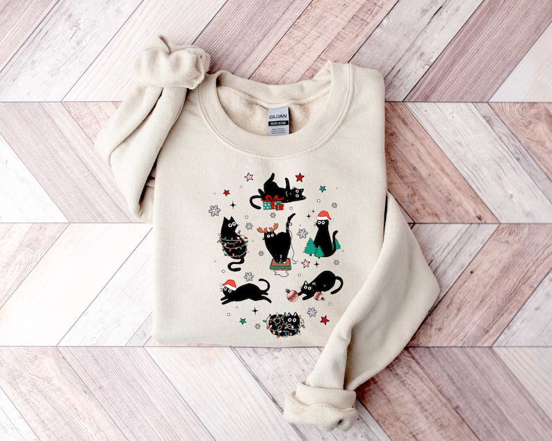 May include: A cream-coloured sweatshirt with a festive design featuring black cats wearing Santa hats, reindeer antlers, and Christmas lights. The cats are surrounded by snowflakes, stars, and Christmas trees.