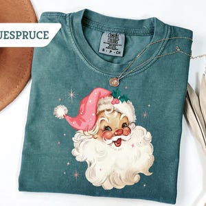 May include: Teal-colored t-shirt featuring a vintage Santa Claus graphic with a pink hat and white beard. The shirt has a crew neck and short sleeves. A gold necklace with a pendant is draped on the shirt. The brand name "BLUESPRUCE" is visible.