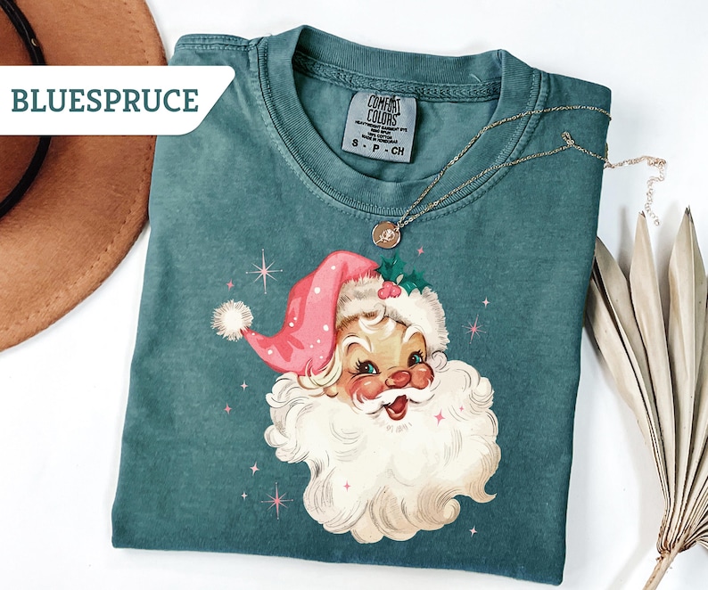 May include: Teal-colored t-shirt featuring a vintage Santa Claus graphic with a pink hat and white beard. The shirt has a crew neck and short sleeves. A gold necklace with a pendant is draped on the shirt. The brand name "BLUESPRUCE" is visible.