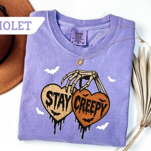May include: Violet-colored t-shirt with a crew neck. The shirt features a skeleton hand holding two heart-shaped pumpkins with the words "Stay Creepy" and dripping black accents. White bat graphics are scattered around the design. A gold necklace is draped on the shirt.