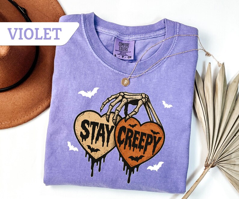 May include: Violet-colored t-shirt with a crew neck. The shirt features a skeleton hand holding two heart-shaped pumpkins with the words "Stay Creepy" and dripping black accents. White bat graphics are scattered around the design. A gold necklace is draped on the shirt.