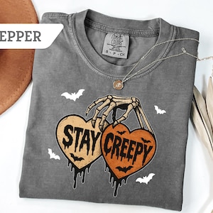 May include: A gray Comfort Colors t-shirt with a Halloween-themed graphic. The graphic features a skeleton hand holding two heart-shaped pumpkins with the words "Stay Creepy". Small white bats are scattered around the design.