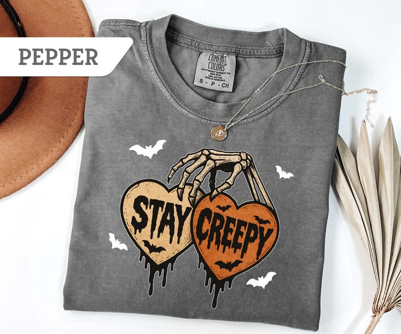 May include: A gray Comfort Colors t-shirt with a Halloween-themed graphic. The graphic features a skeleton hand holding two heart-shaped pumpkins with the words "Stay Creepy". Small white bats are scattered around the design.