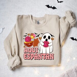 May include: A beige sweatshirt featuring a ghost holding a pink and white pastry. The text "Aqui Espantan" is printed on the sweatshirt.