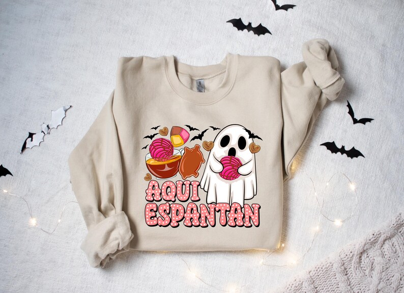 May include: A beige sweatshirt featuring a ghost holding a pink and white pastry. The text "Aqui Espantan" is printed on the sweatshirt.