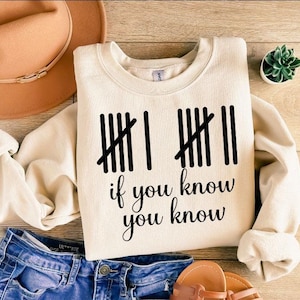 May include: Cream-colored sweatshirt with the phrase "if you know you know" in black script. The design includes tally marks. The sweatshirt is paired with blue denim shorts, a brown hat, sandals, and a small succulent plant.