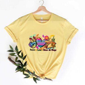 Puede incluir: Camiseta amarilla con un s&iacute;mbolo de paz, un coraz&oacute;n y un sombrero. El texto "Peace Love Cinco de Mayo" est&aacute; impreso debajo del gr&aacute;fico.