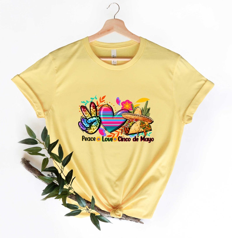 Puede incluir: Camiseta amarilla con un s&iacute;mbolo de paz, un coraz&oacute;n y un sombrero. El texto "Peace Love Cinco de Mayo" est&aacute; impreso debajo del gr&aacute;fico.