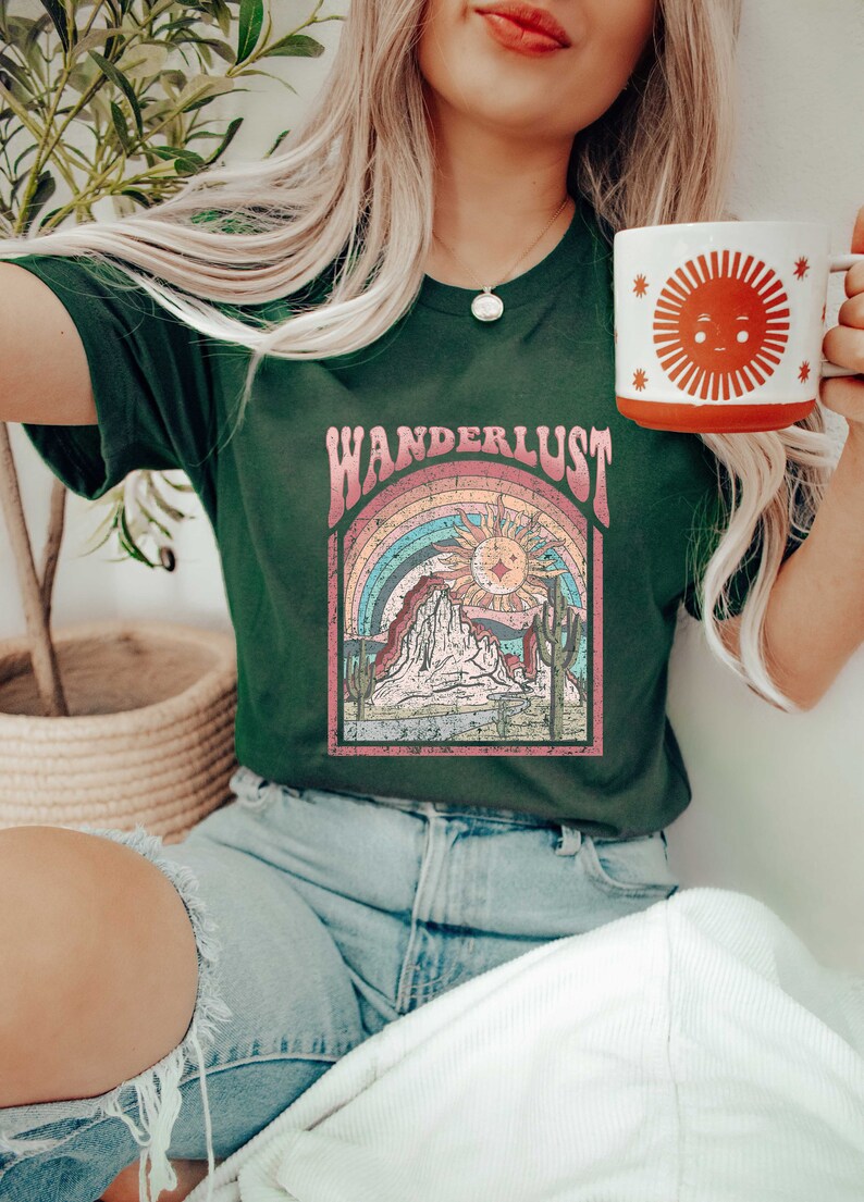 May include: A green t-shirt with a vintage-style graphic of a mountain range, sun, and rainbow. The text "WANDERLUST" is printed in pink and white letters above the graphic.