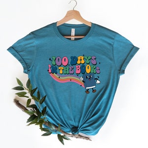 May include: Teal blue t-shirt with a rainbow and stars graphic. The graphic says "100 Days in the Books" and features a cartoon book character.