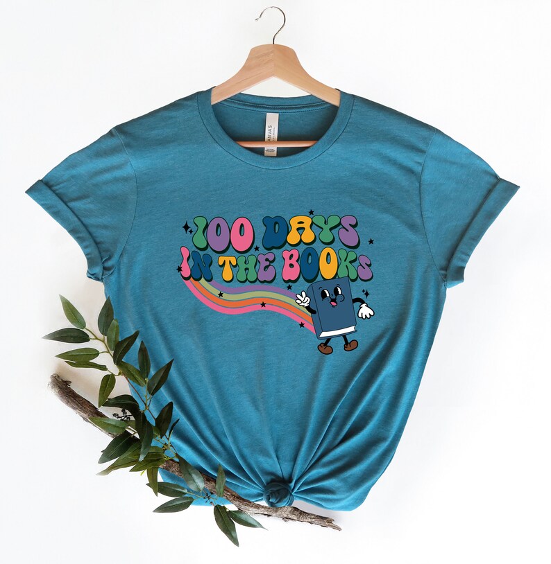 May include: Teal blue t-shirt with a rainbow and stars graphic. The graphic says "100 Days in the Books" and features a cartoon book character.