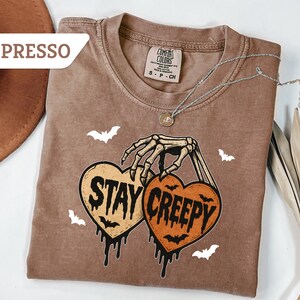 May include: Espresso-colored t-shirt with a skeleton hand holding two heart-shaped pumpkins that read "Stay Creepy." The shirt has a crew neck and short sleeves. White bat graphics are scattered around the design. A silver necklace with a pendant is draped across the shirt.