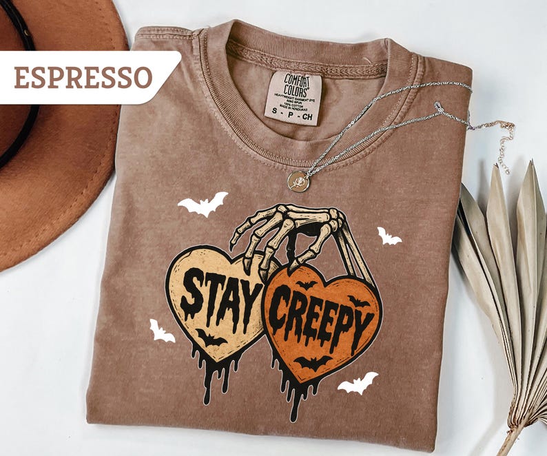 May include: Espresso-colored t-shirt with a skeleton hand holding two heart-shaped pumpkins that read "Stay Creepy." The shirt has a crew neck and short sleeves. White bat graphics are scattered around the design. A silver necklace with a pendant is draped across the shirt.