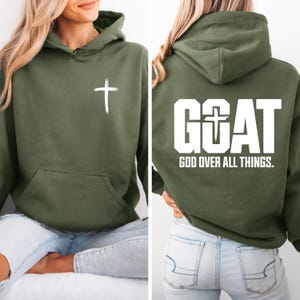 Pode incluir: Moletom verde-oliva com uma cruz branca na frente e o texto "GOAT GOD OVER ALL THINGS" nas costas. Possui capuz e bolso frontal. Feito de um material macio.