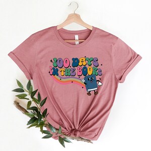 May include: A pink t-shirt with a rainbow and a cartoon book character. The text on the shirt reads "100 Days in the Books".