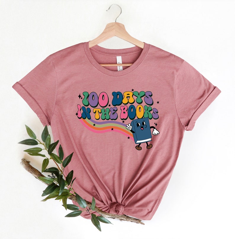 May include: A pink t-shirt with a rainbow and a cartoon book character. The text on the shirt reads "100 Days in the Books".