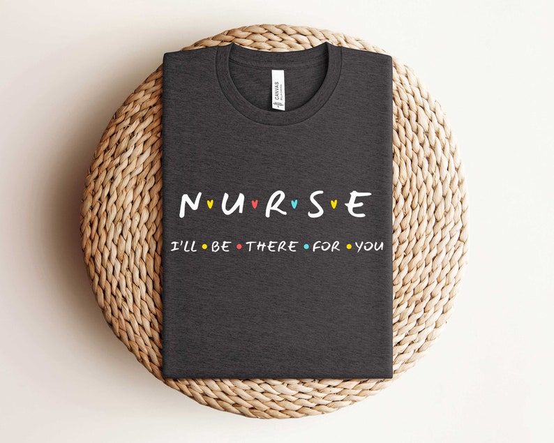 Puede incluir: Camiseta gris oscuro con el texto "NURSE" en letras de colores con corazones entre cada letra. Debajo del texto est&aacute; "I'll be there for you" en letras negras con peque&ntilde;os c&iacute;rculos entre cada palabra.