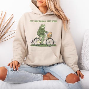 May include: A beige hoodie with the text "OFF TO BE RADICAL LEFT SCUM" above a cartoon frog riding a bicycle with a cat in the basket. The frog wears a green sweater. The bicycle is white with black tires.