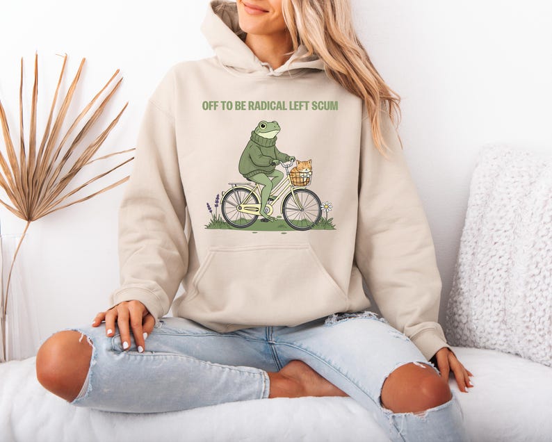 May include: A beige hoodie with the text "OFF TO BE RADICAL LEFT SCUM" above a cartoon frog riding a bicycle with a cat in the basket. The frog wears a green sweater. The bicycle is white with black tires.