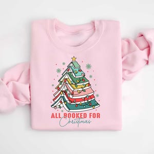 May include: A light pink sweatshirt with a Christmas tree design made of books. The tree is decorated with colorful lights and topped with a gold star. The text "ALL BOOKED FOR Christmas" is printed below the tree.