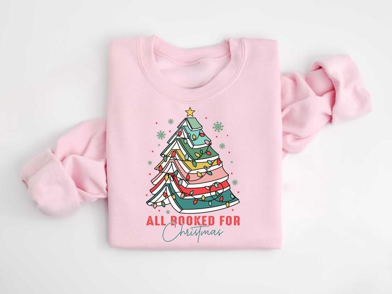 May include: A light pink sweatshirt with a Christmas tree design made of books. The tree is decorated with colorful lights and topped with a gold star. The text "ALL BOOKED FOR Christmas" is printed below the tree.
